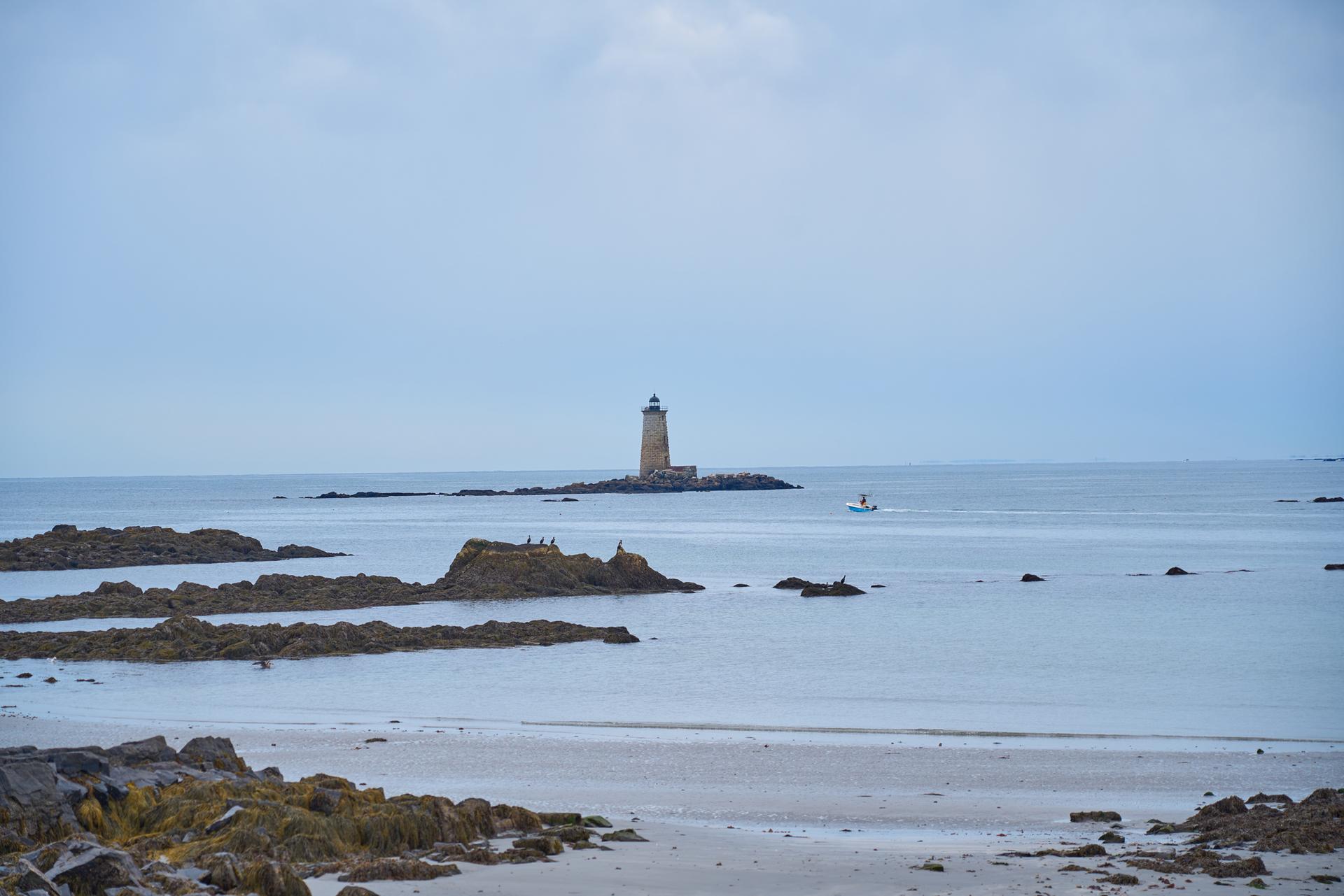 Distant lighthouse on a rocky island in a calm, overcast ocean.
