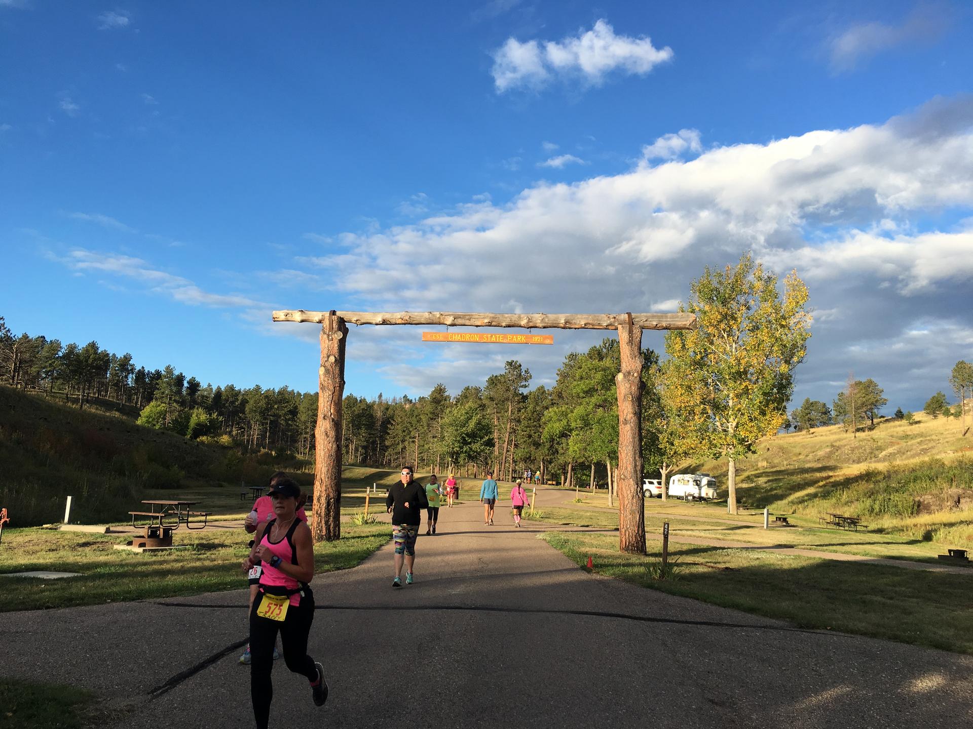 Runners pass under a rustic wooden archway on a sunny day.