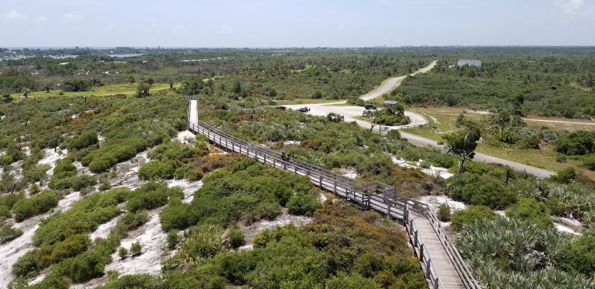 An aerial view of a long wooden boardwalk winding through a green, scrubby landscape.