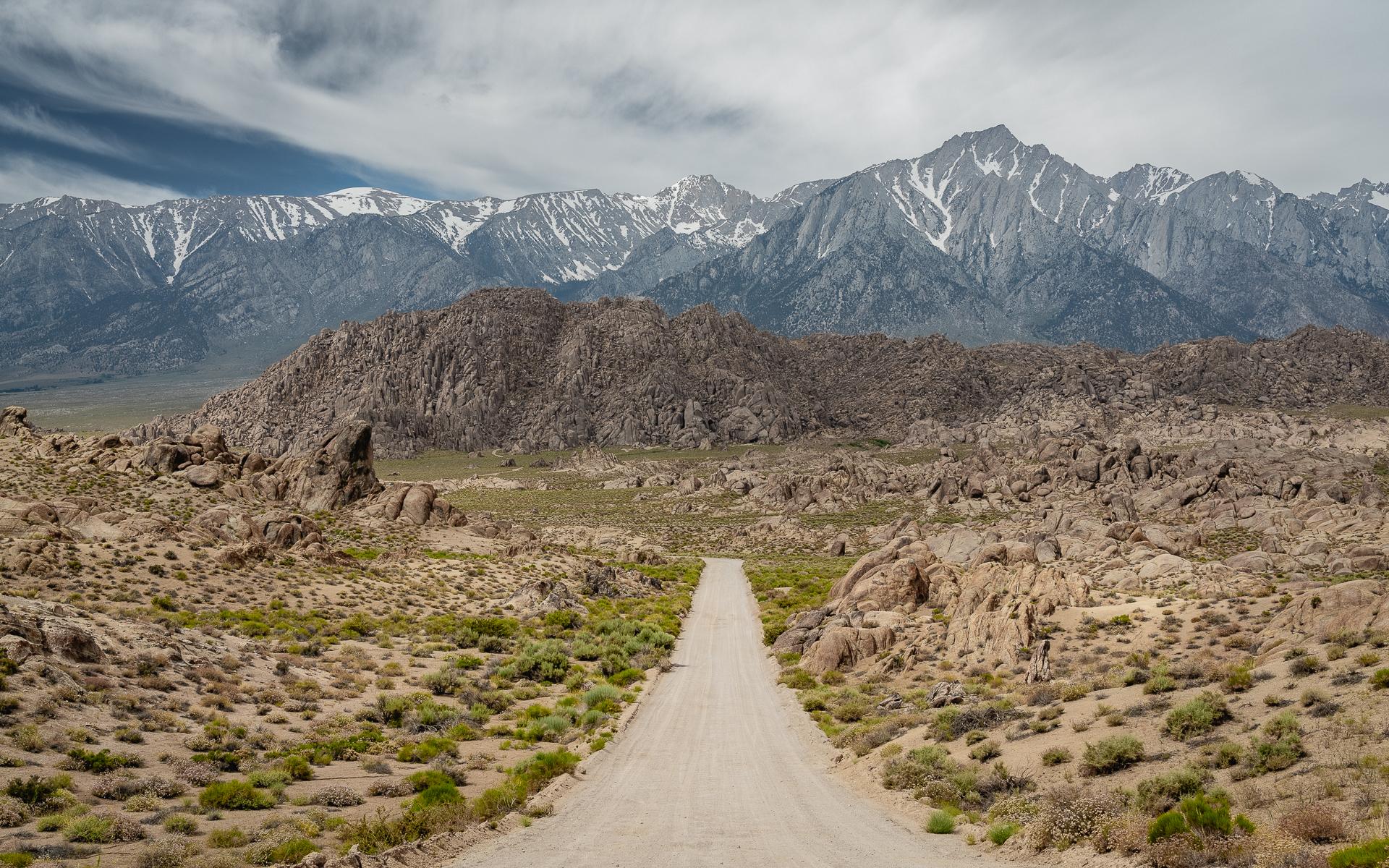 A long dirt road stretches through a rocky desert towards distant snowy mountains.