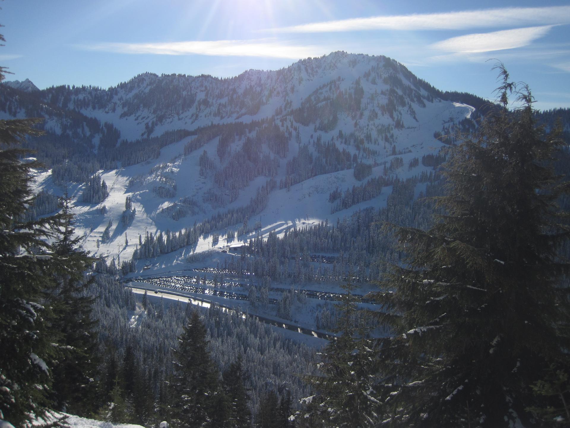 Snow-covered mountains with ski slopes, evergreen trees, and a sunny parking lot.
