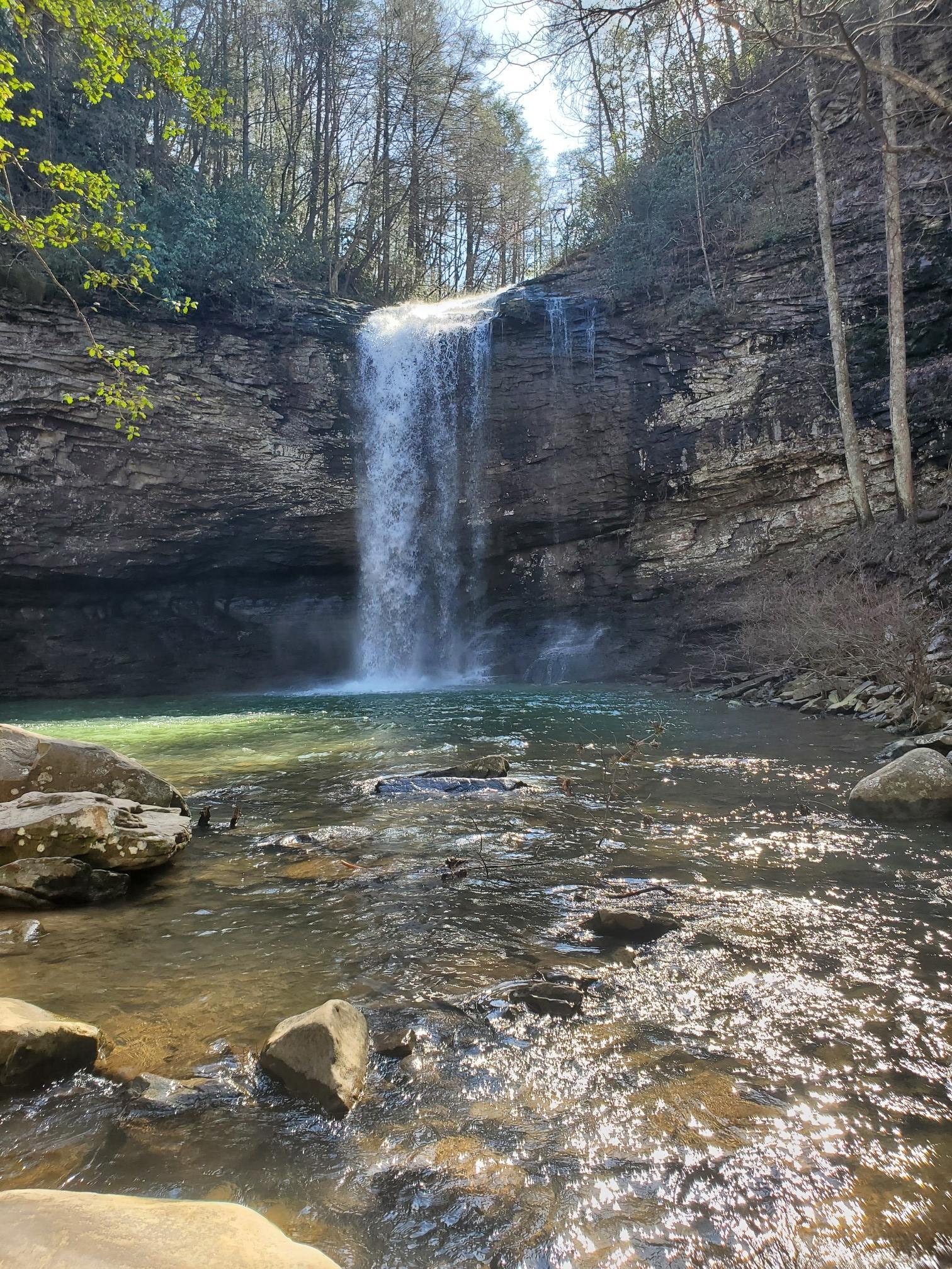 A wide waterfall plunges into a clear pool, with a sunlit stream.
