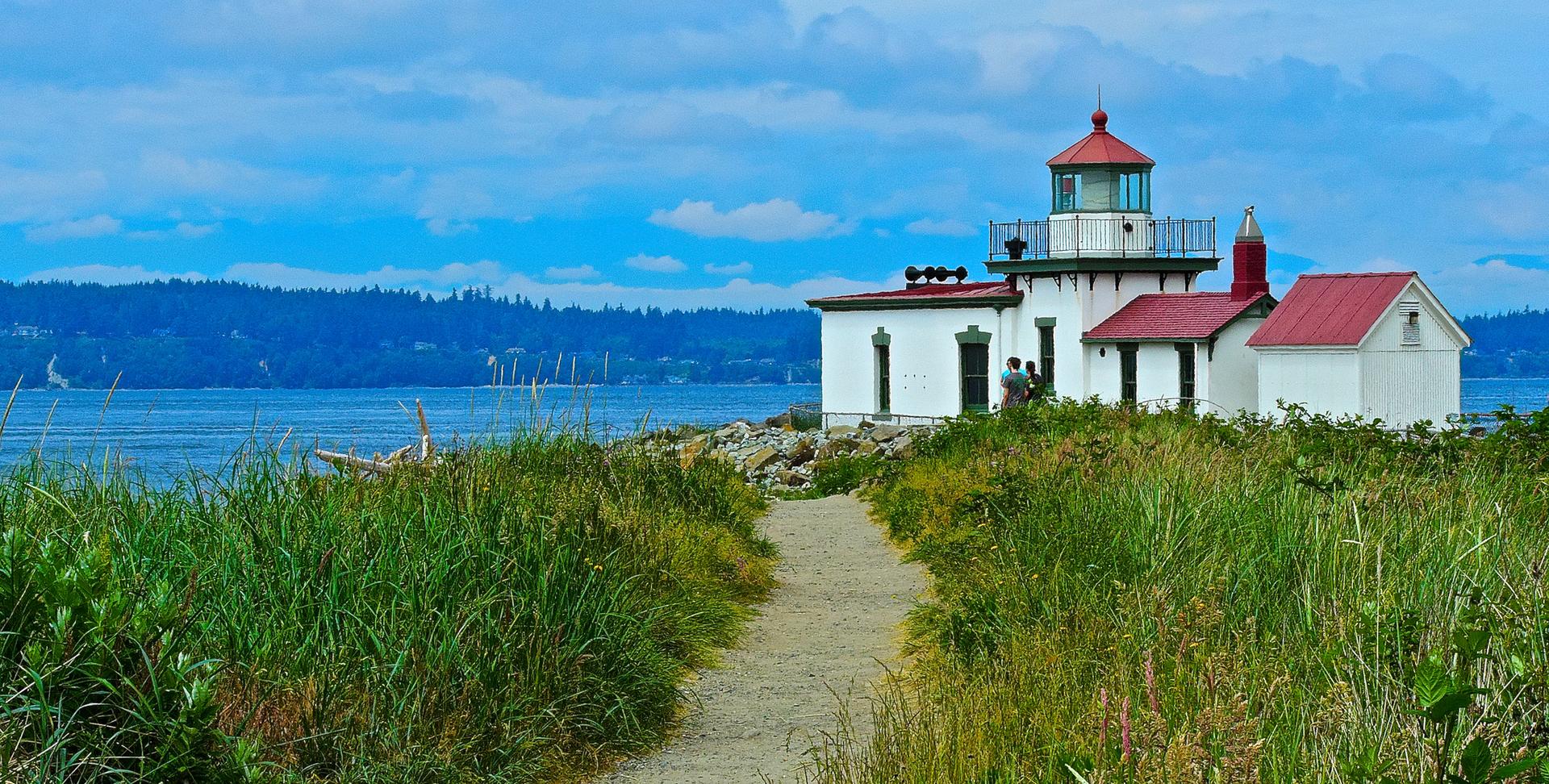 White lighthouse with red roof by blue water. Grassy path leads to the structure.