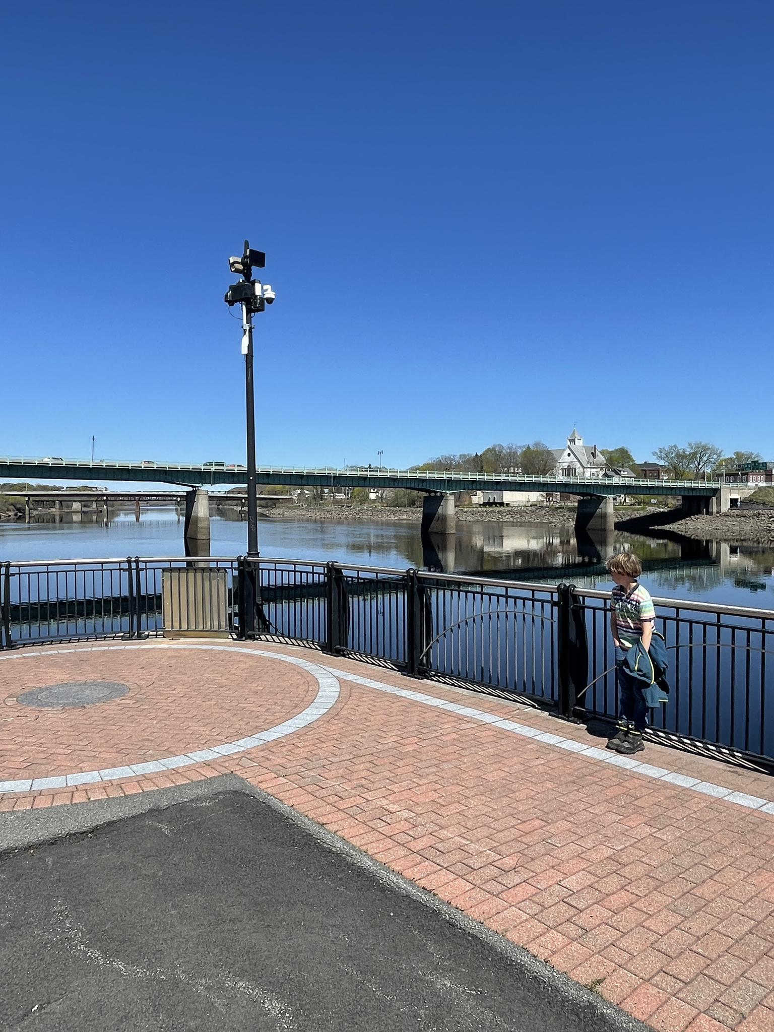 Person fishing from a promenade overlooking a river and distant bridge.