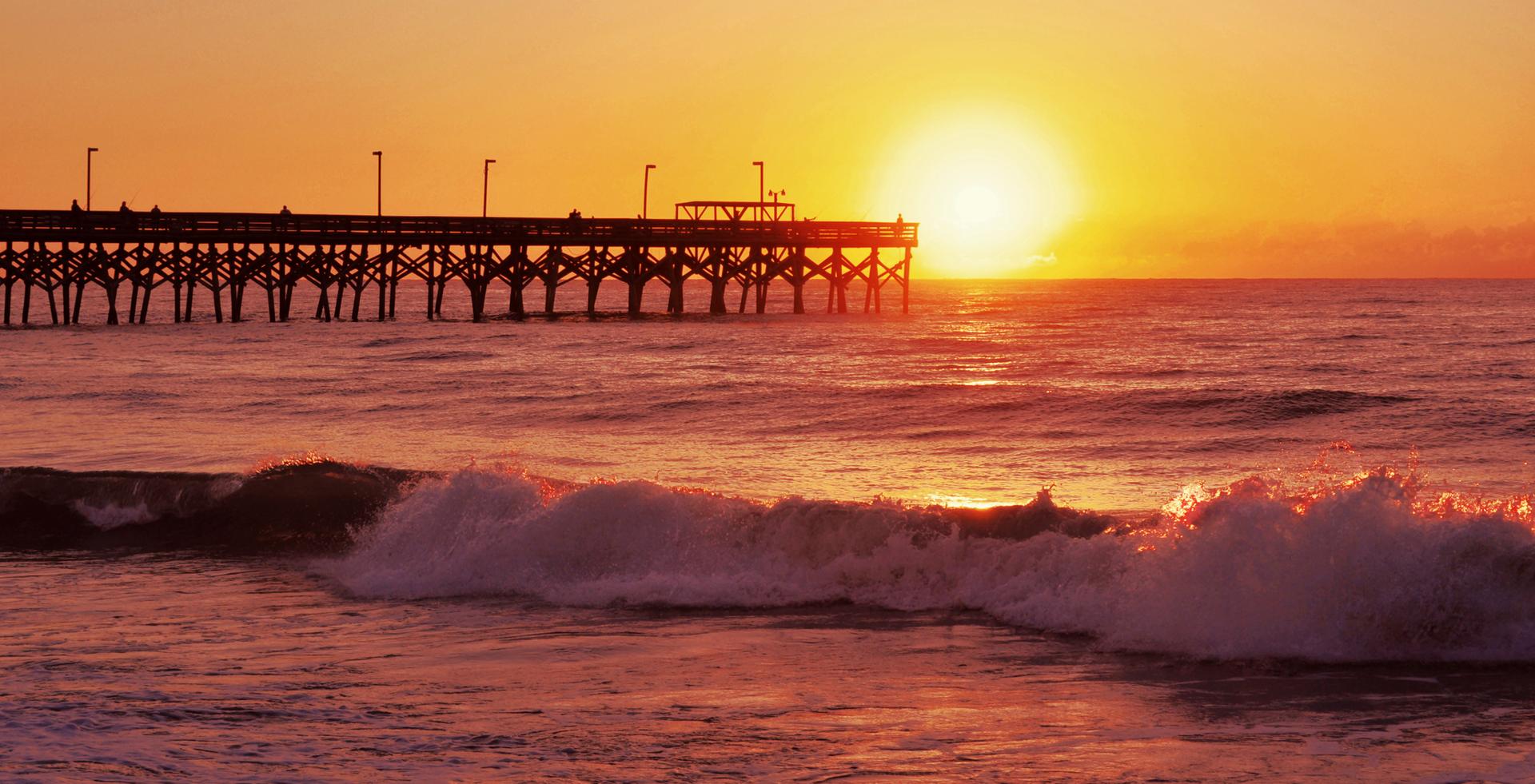 Golden sunrise illuminates ocean waves and a long wooden pier.