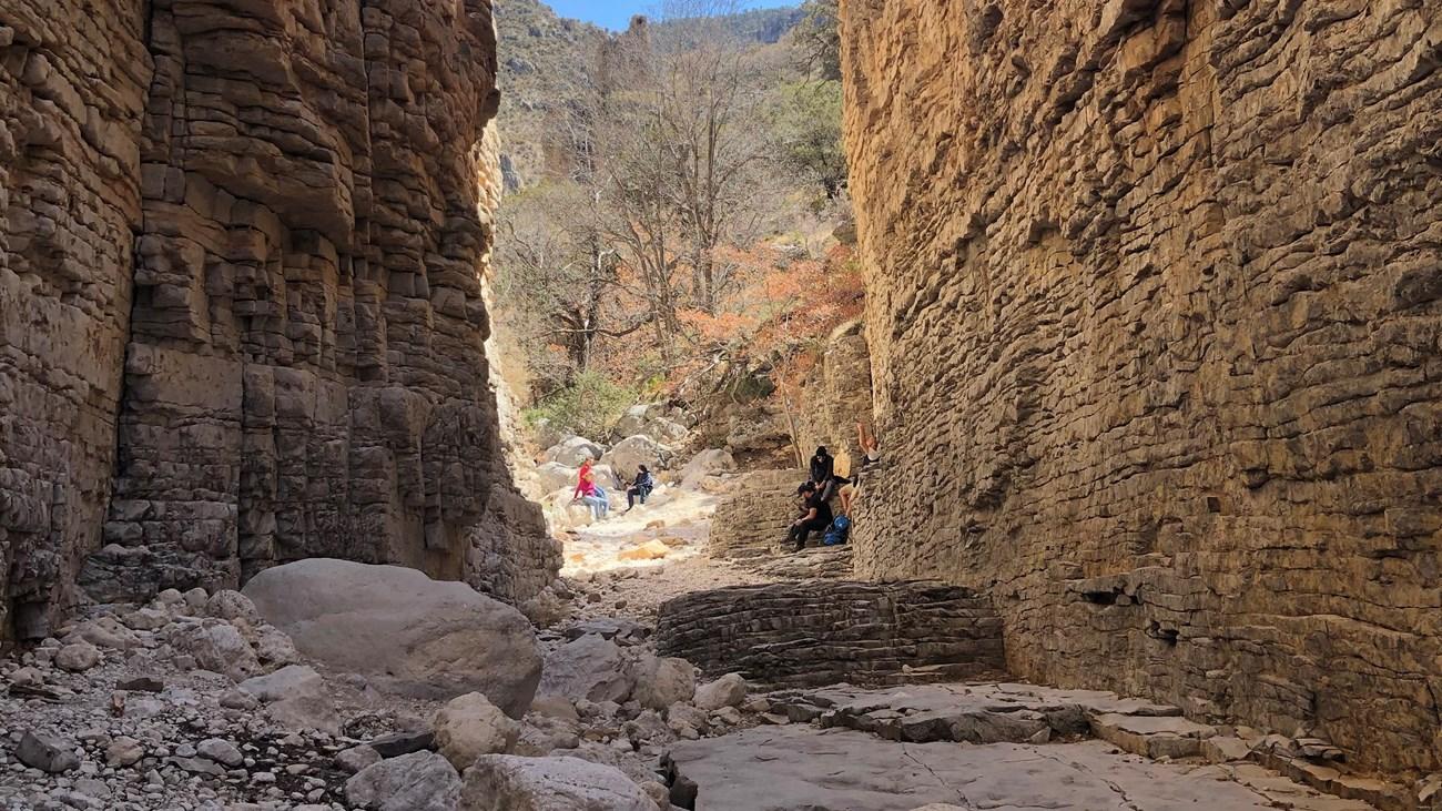 Hikers in a narrow canyon with tall, layered rock walls and dry streambed.