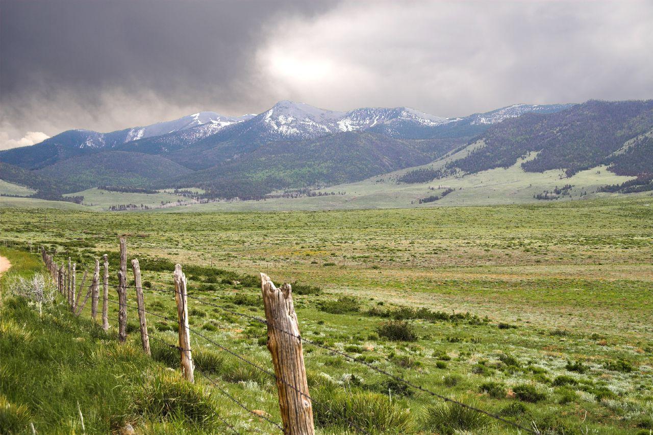 A rustic fence line in a green field with snow-capped mountains under cloudy skies.