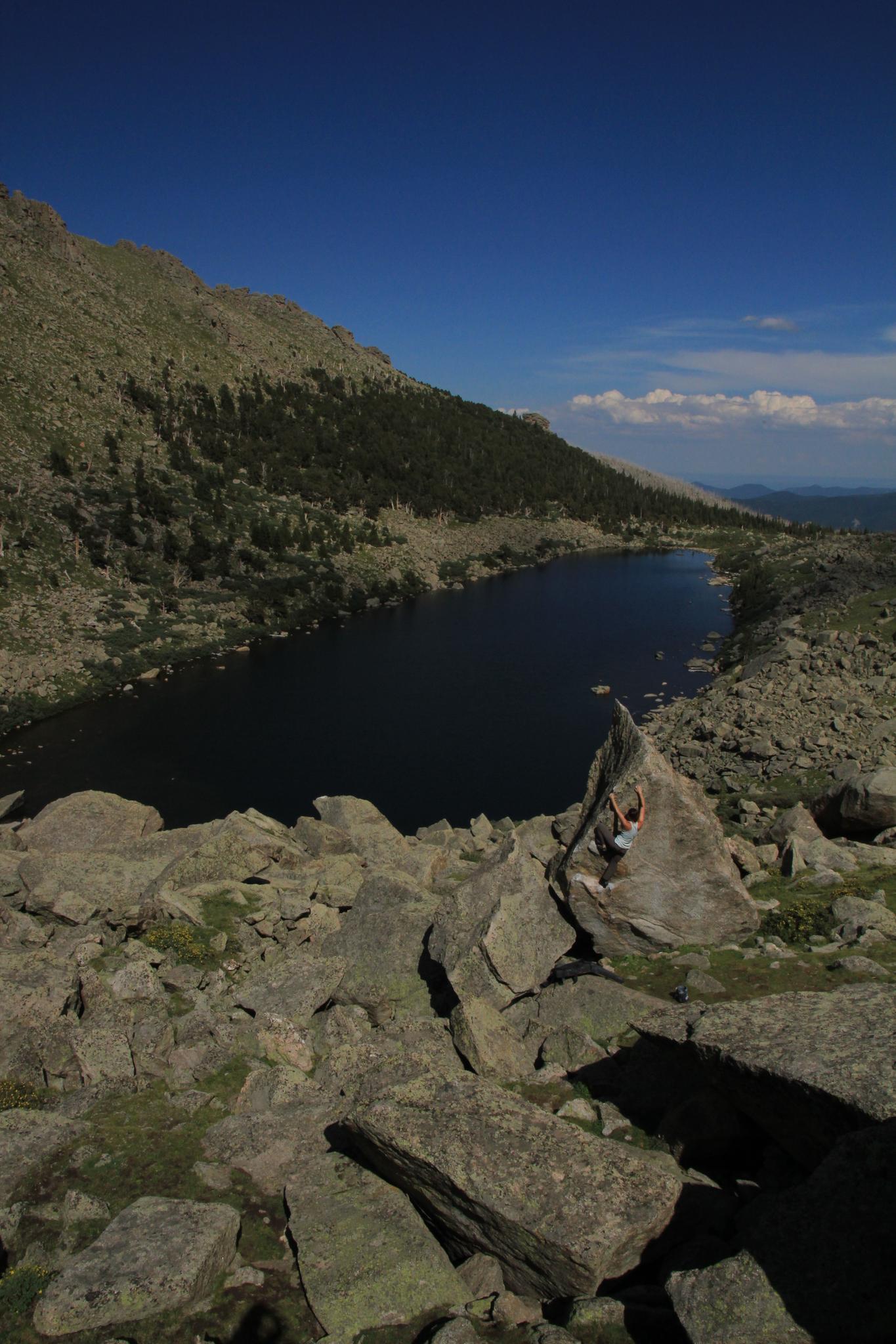 Person sits on rocky outcrop overlooking a dark mountain lake under bright blue sky.