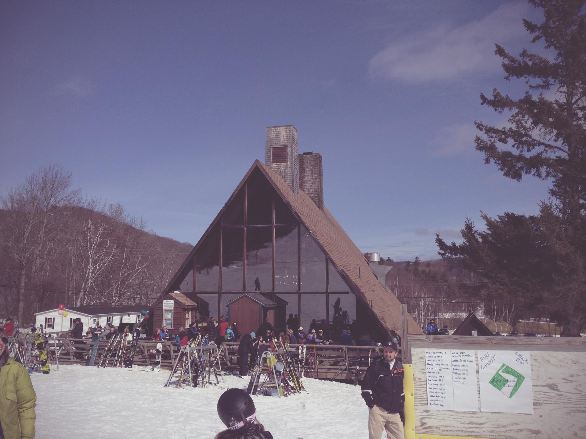 People gather outside a large A-frame lodge on a sunny, snowy day.
