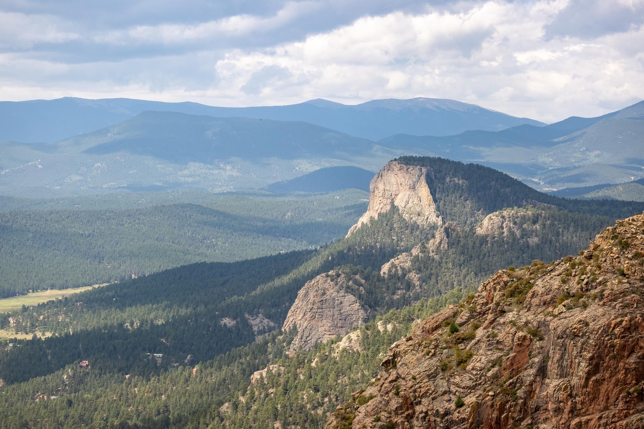 Panoramic view of rugged mountains, forested valleys, and rocky peaks under a cloudy sky.