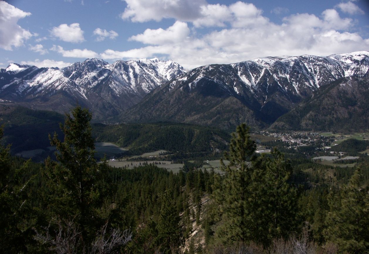Snowy mountains overlook a forested valley and distant town under a cloudy sky. - Sauer's Mountain Trail