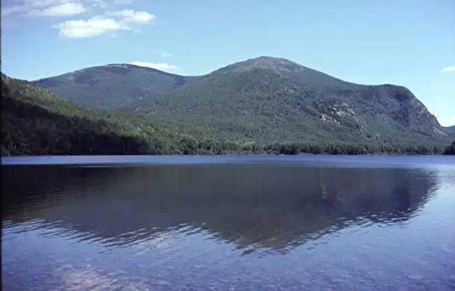 Calm lake reflecting tree-covered mountains under a bright blue sky.