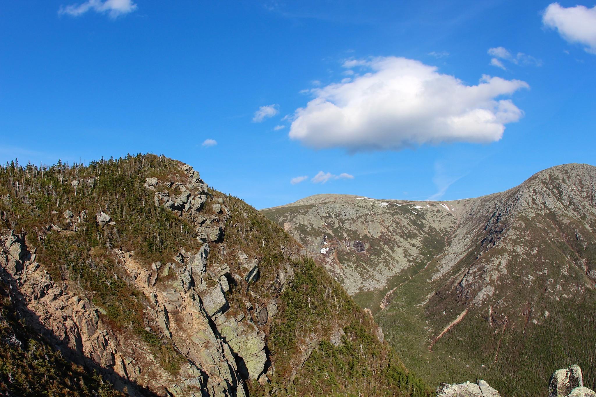 Rugged mountain landscape with rocky peaks, green trees, and a bright blue sky.