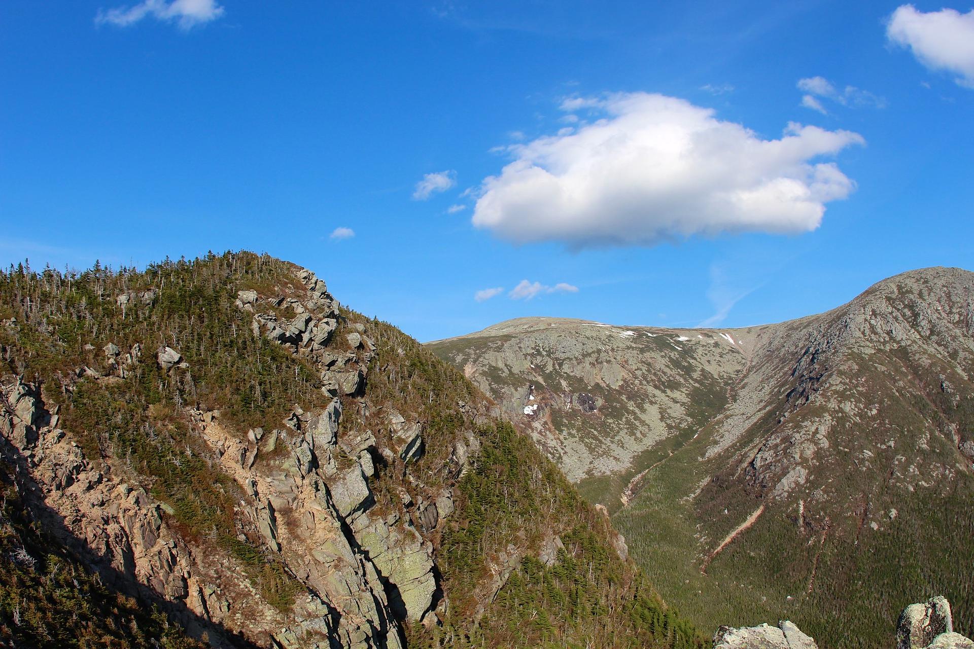 Rugged mountain landscape with rocky peaks, green trees, and a bright blue sky.