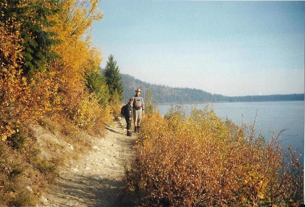 Two hikers on a dirt path beside a lake, surrounded by autumn foliage.