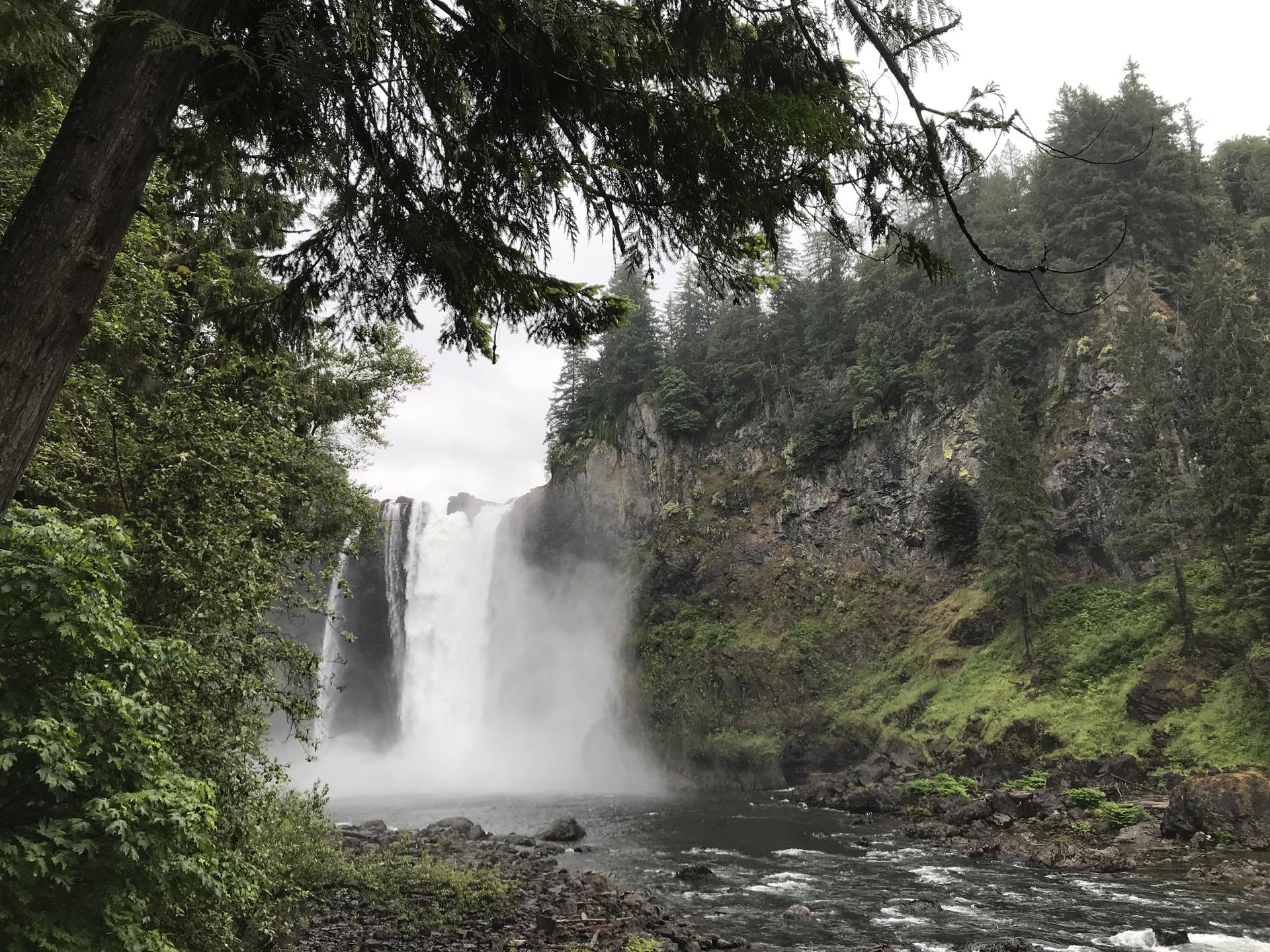 A powerful waterfall plunges into a river, surrounded by rocky cliffs and green trees.