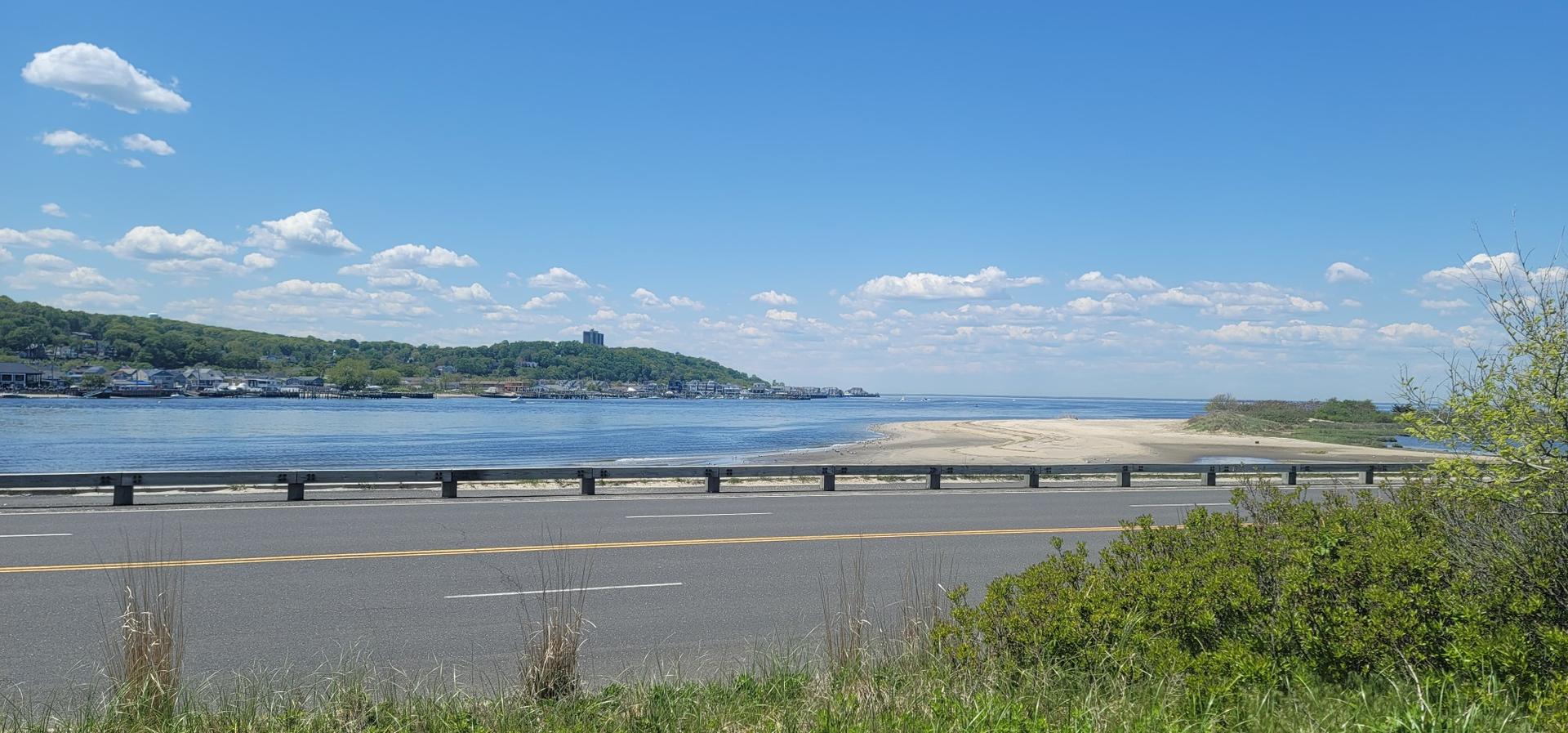 Coastal road next to calm blue water, sandy beach, and distant green hills.