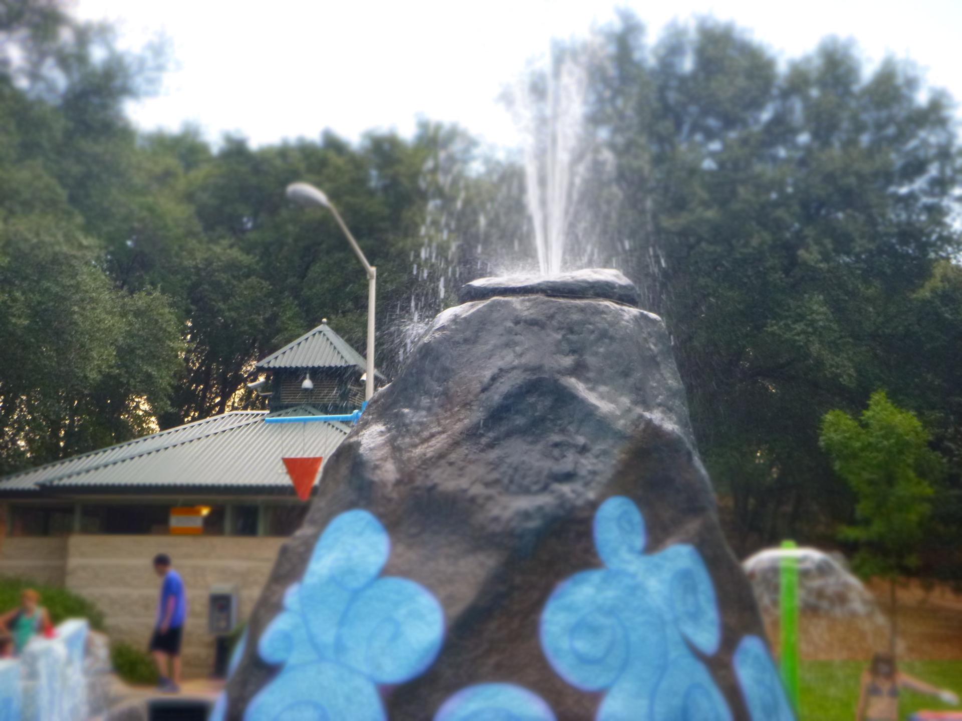 Water sprays from a rock fountain with blue designs, people and trees visible.