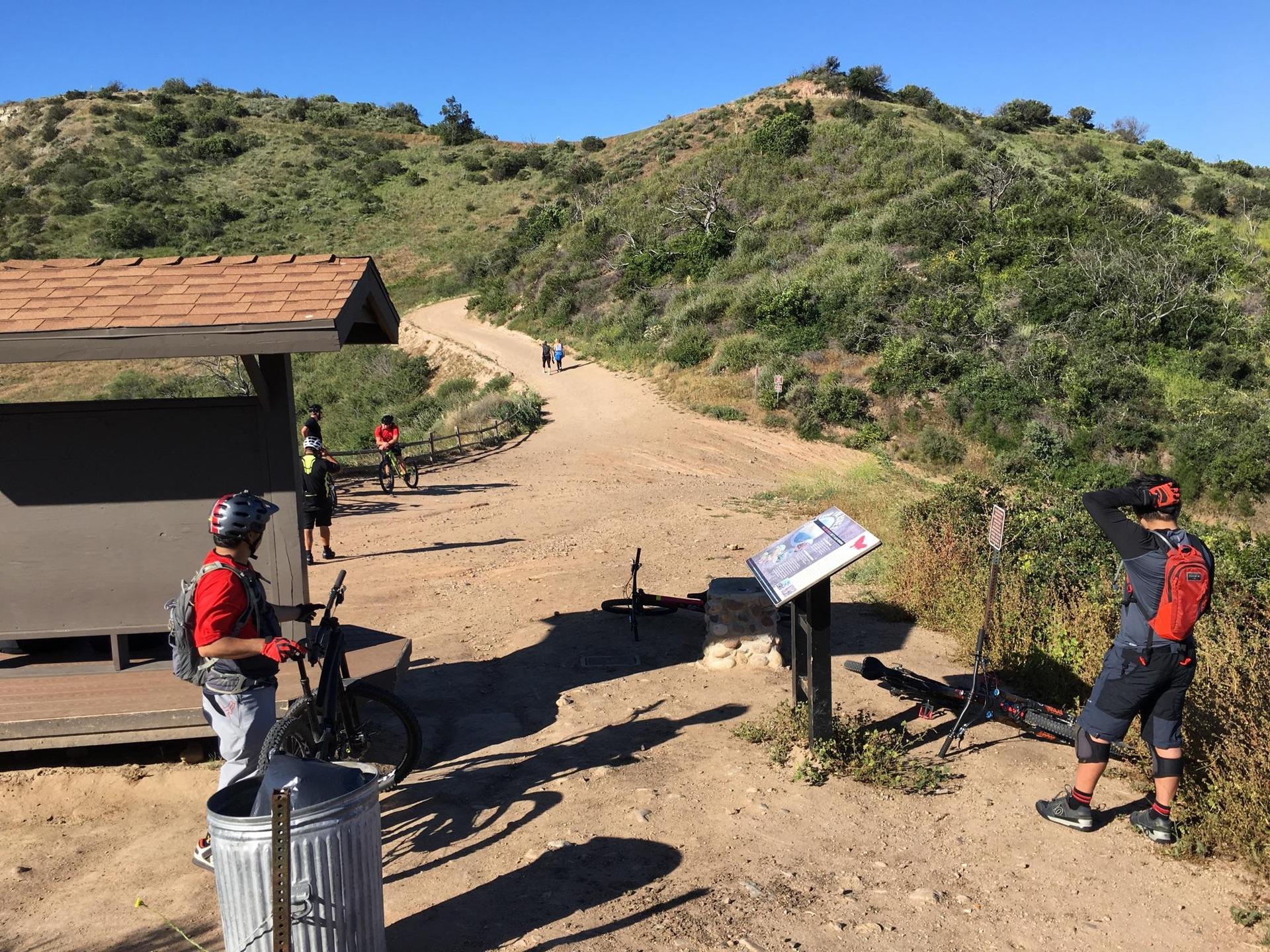 Mountain bikers gathered at a trail junction with a shelter on a sunny day.