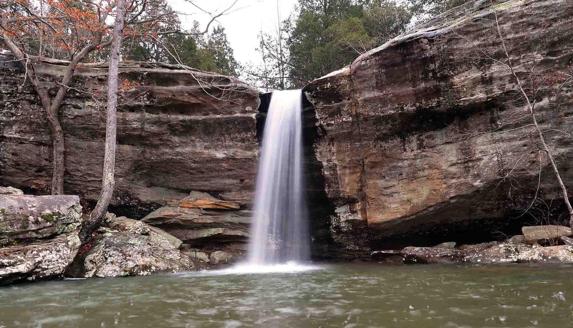 A serene waterfall flows over layered rock formations into a dark pool.