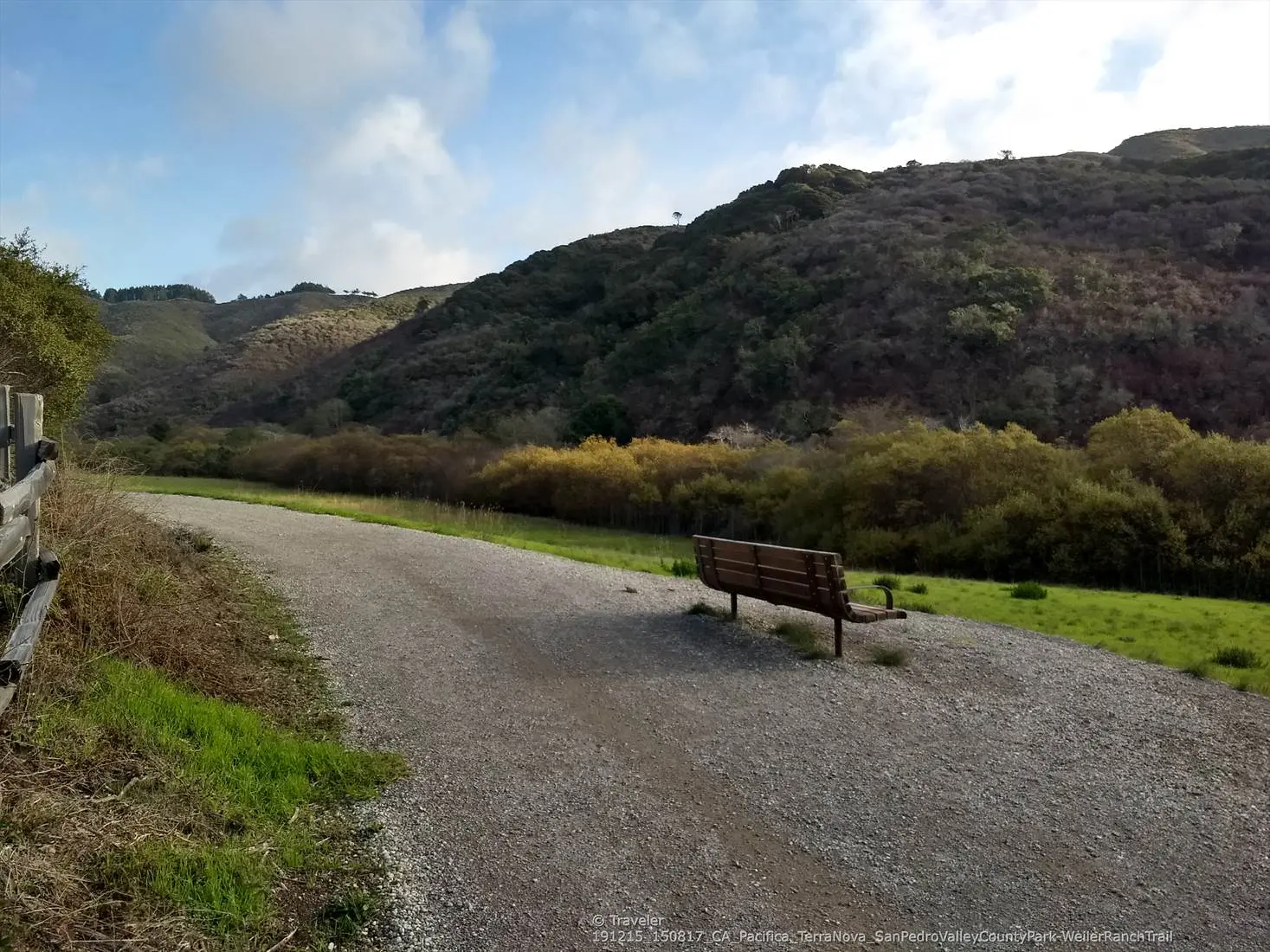 Gravel path with a wooden bench, facing hills and green foliage. Bright sky.