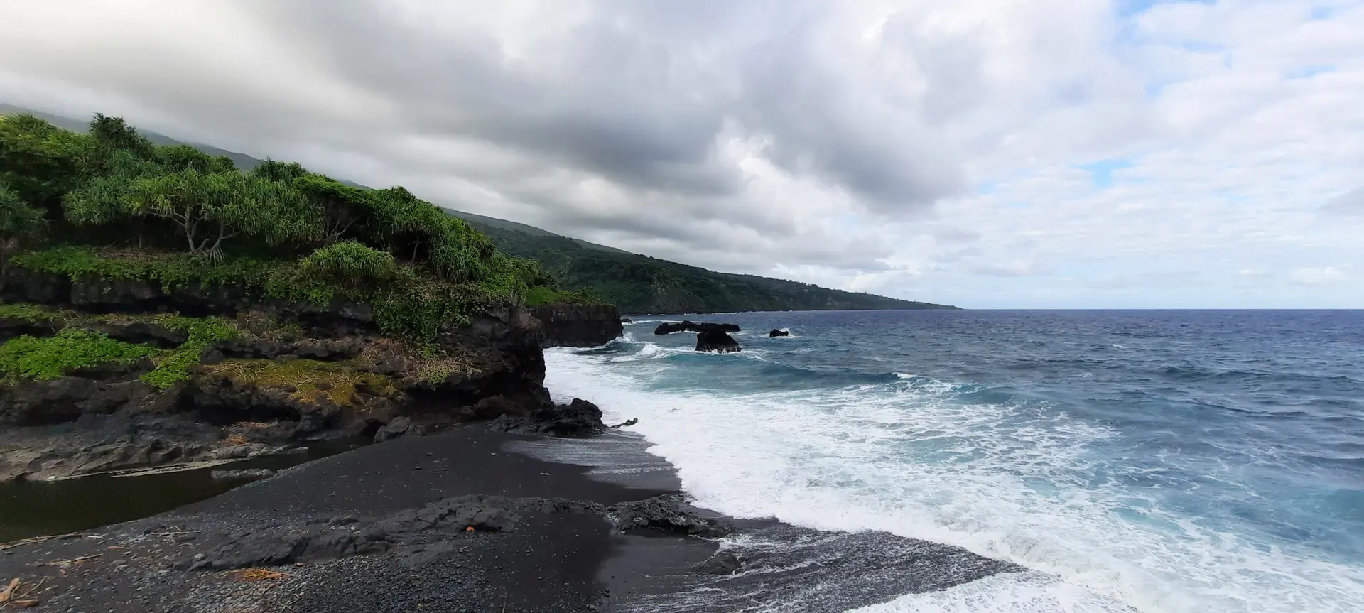 Panoramic view of dark sand beach, crashing waves, and green cliffs.