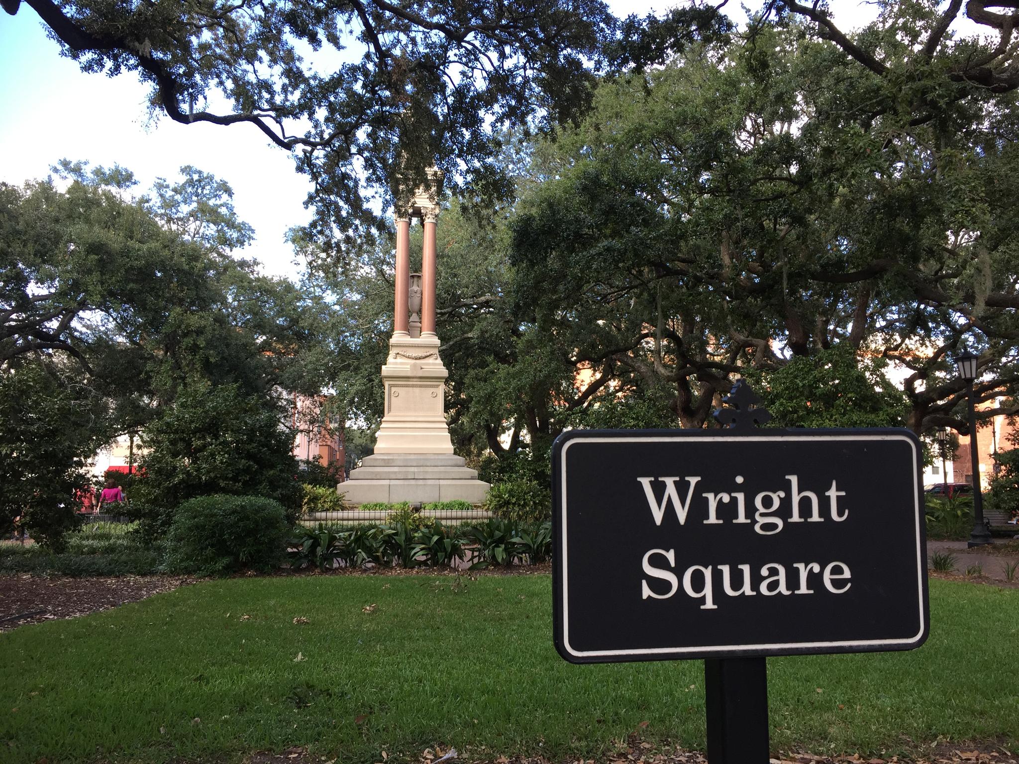 A monument in a grassy square with large trees and a black sign.