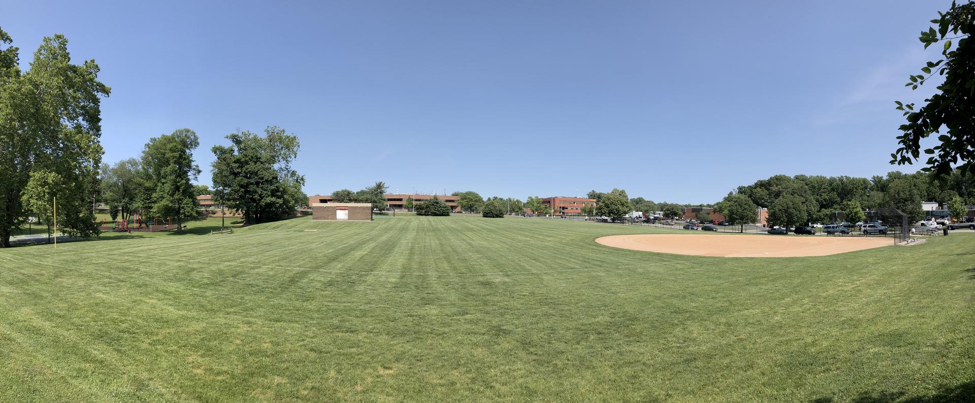 Expansive green field with a baseball diamond under a clear, sunny sky.