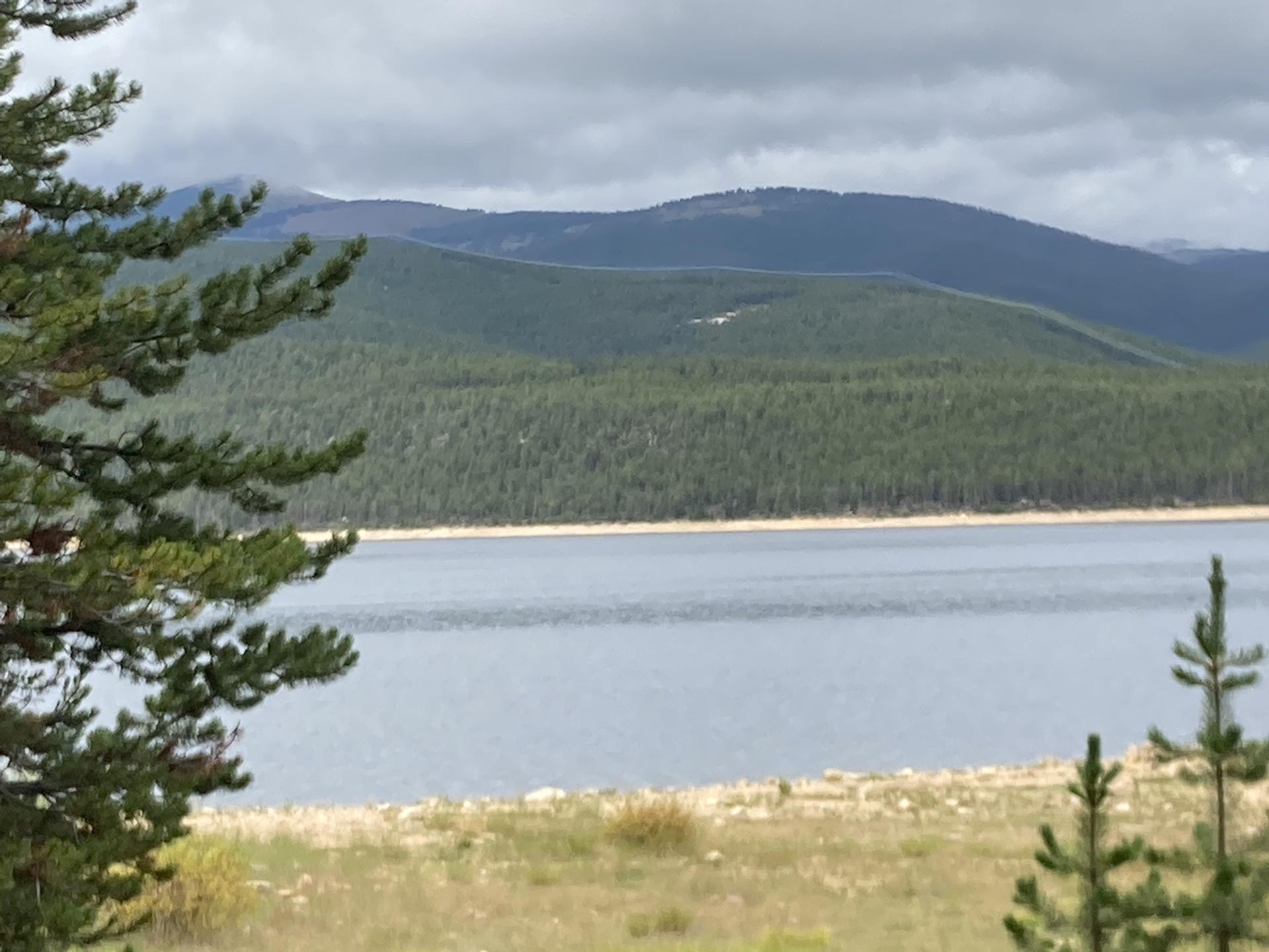 Calm lake with forested hills and distant mountains under a cloudy sky.