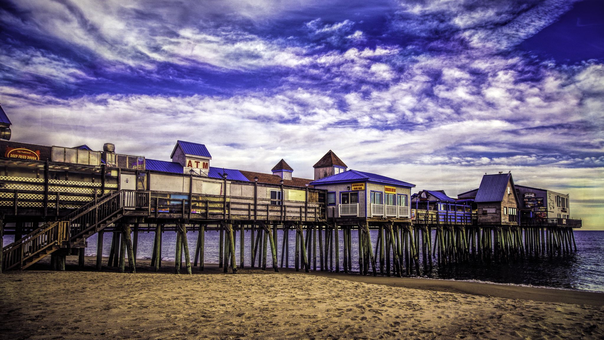 Wooden pier with buildings over water, sandy beach, and dramatic blue sky. - Eastern Trail
