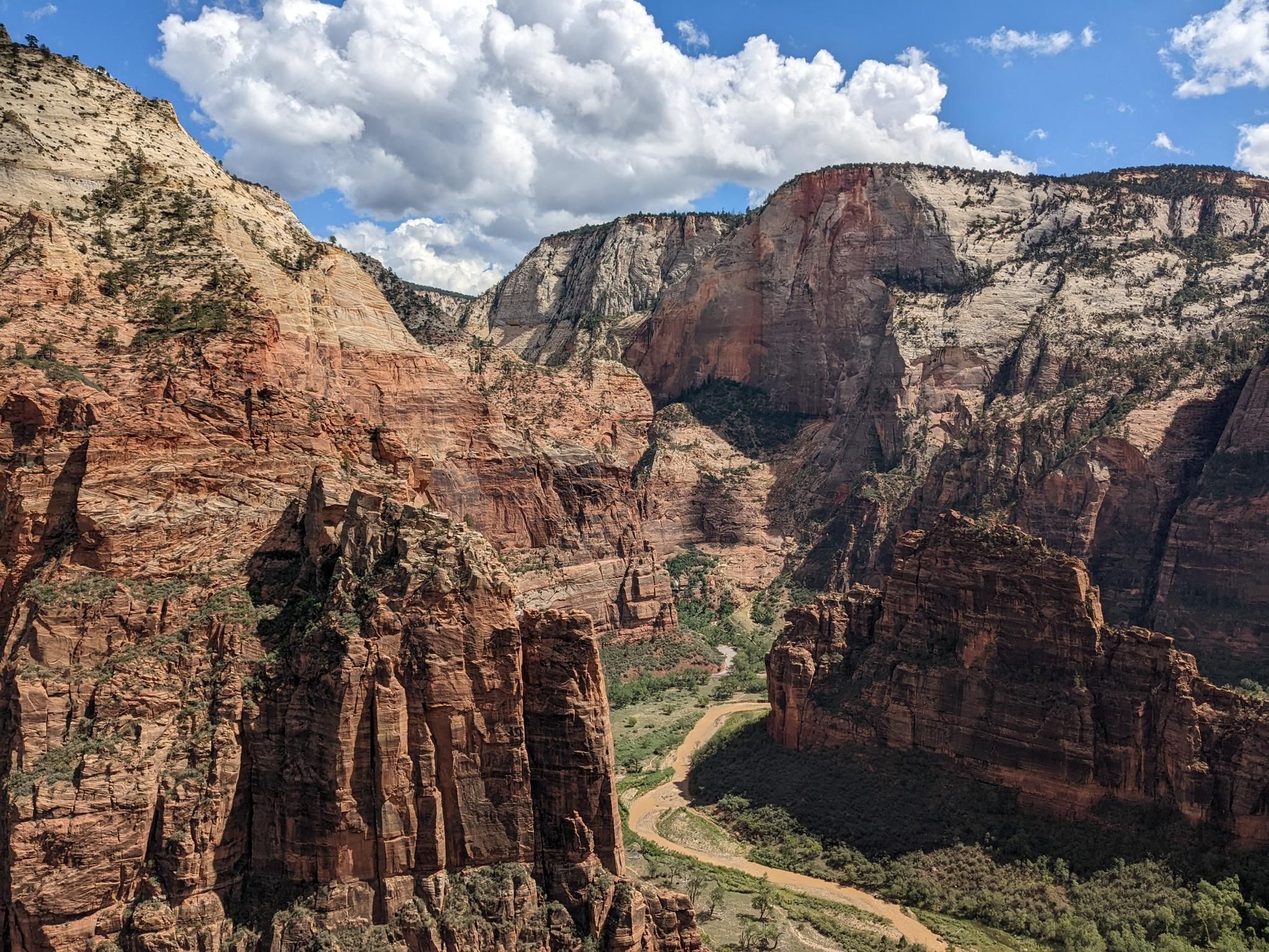 Vast canyon landscape with towering red cliffs, a winding river, and cloudy sky. - Angels Landing Trail