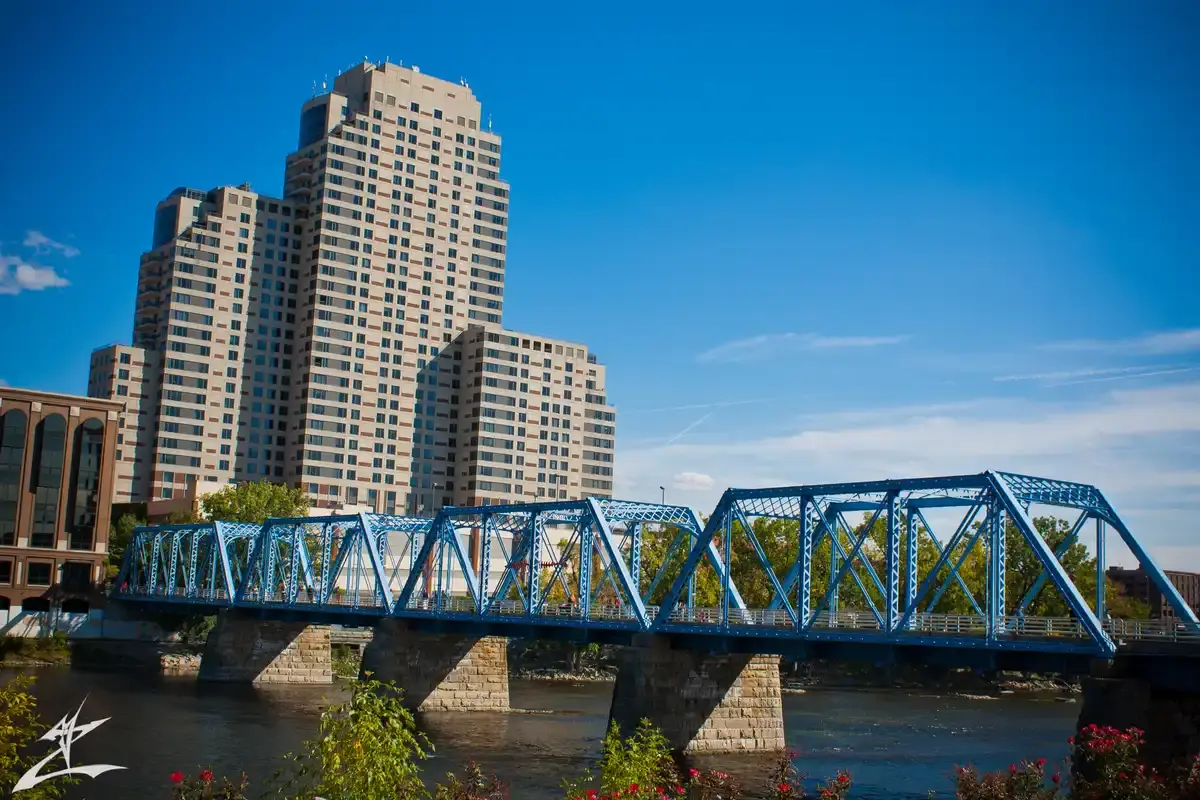 Blue truss bridge over a river with tall buildings under a bright sky.