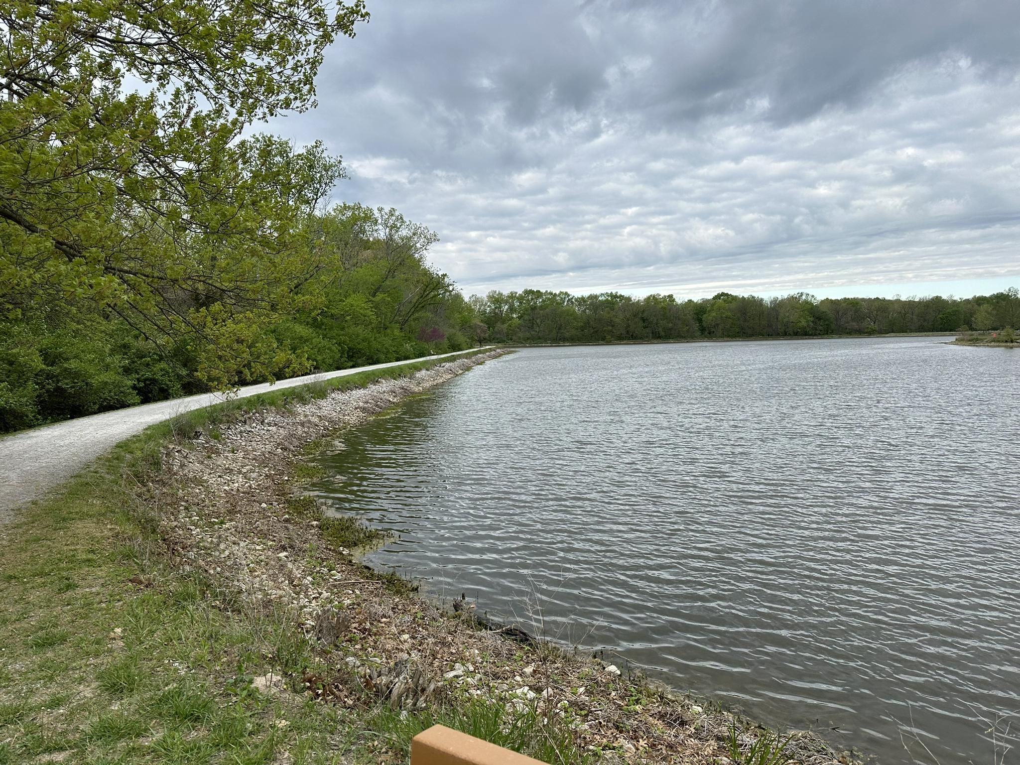 A gravel path curves along a lake under a cloudy sky, surrounded by green trees. - Fishing Lake – MKT Trail loop from Rejoice Free Will Baptist Church
