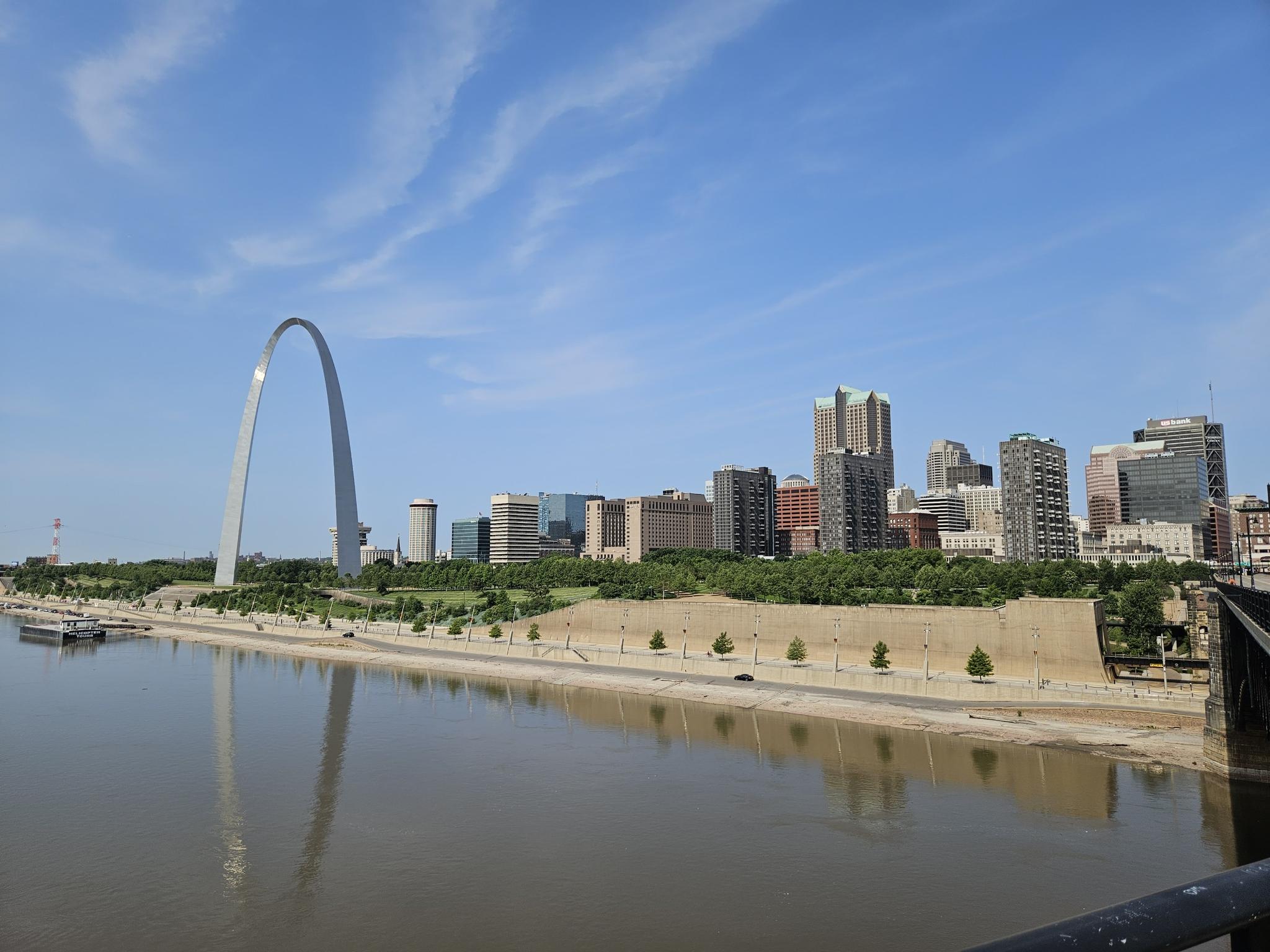 Large arch and city skyline reflected in a wide river under a blue sky.