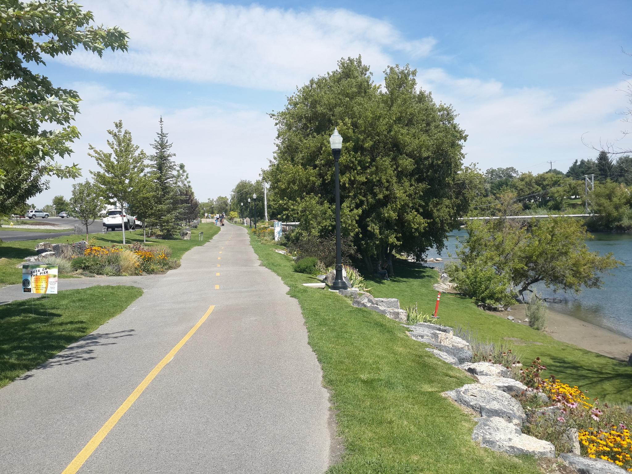 Paved path next to a river, with green grass, trees, and bright sunshine.