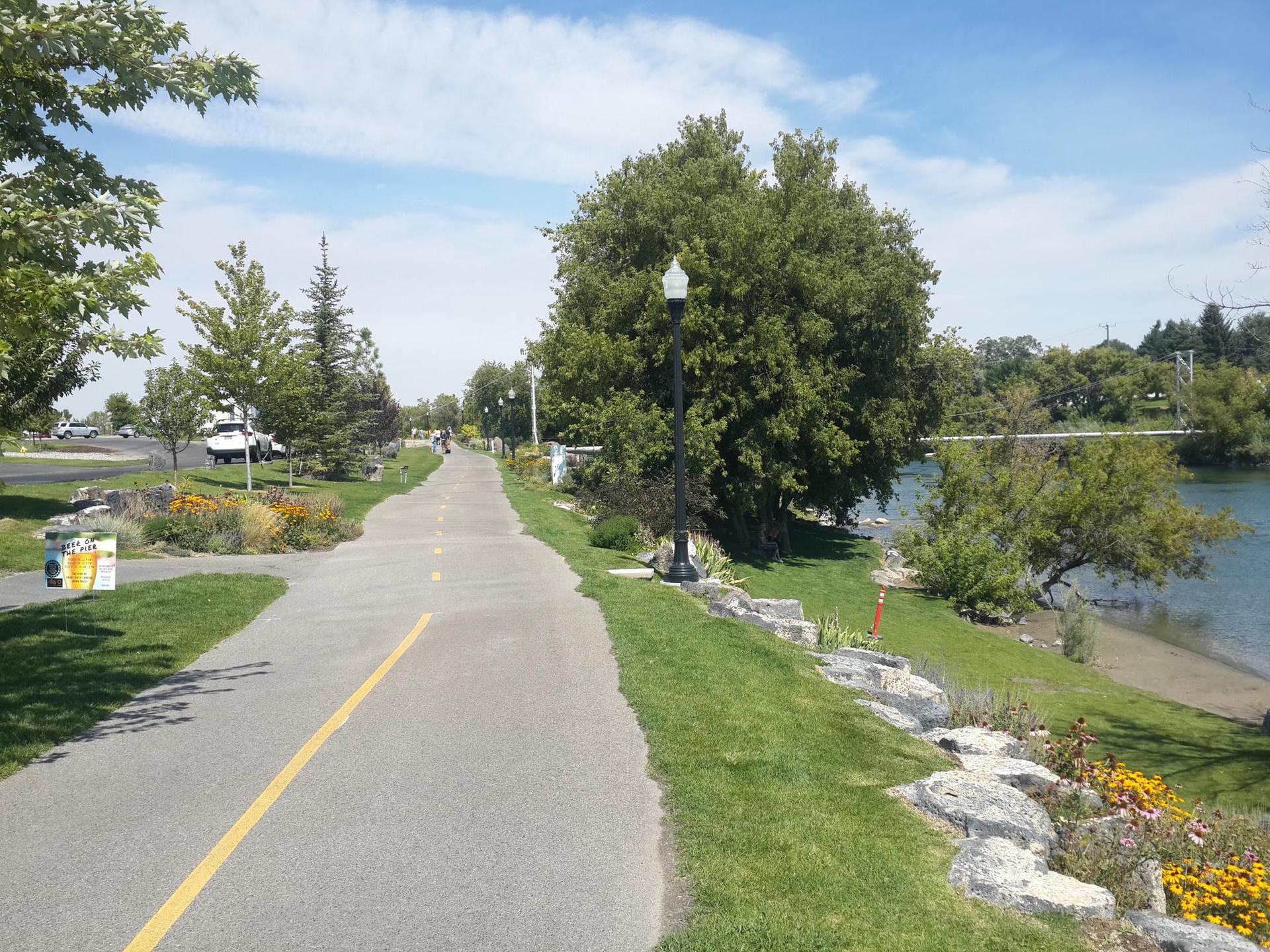 Paved path next to a river, with green grass, trees, and bright sunshine.