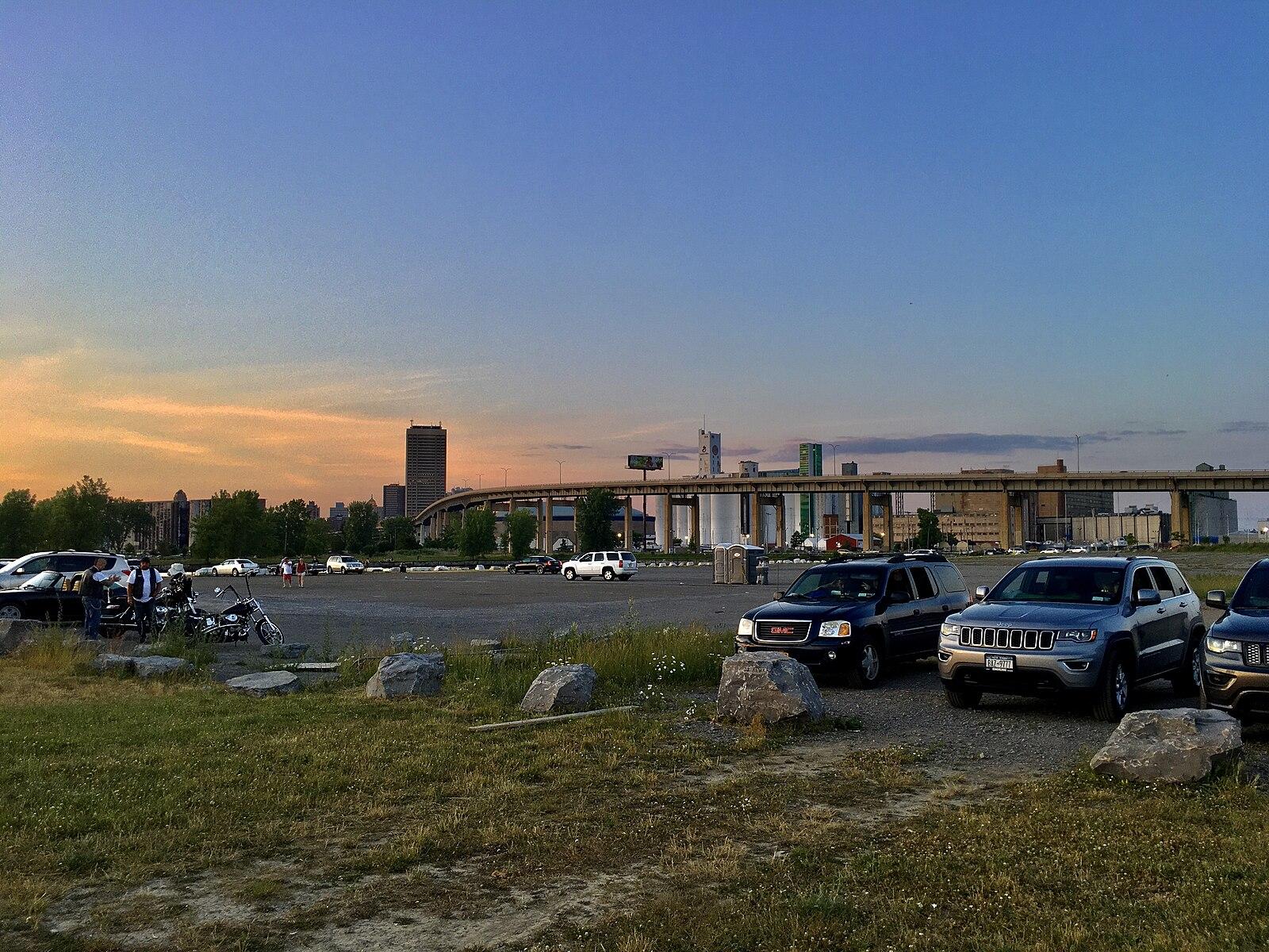 Parking lot at dusk with cars, urban skyline, and distant people.