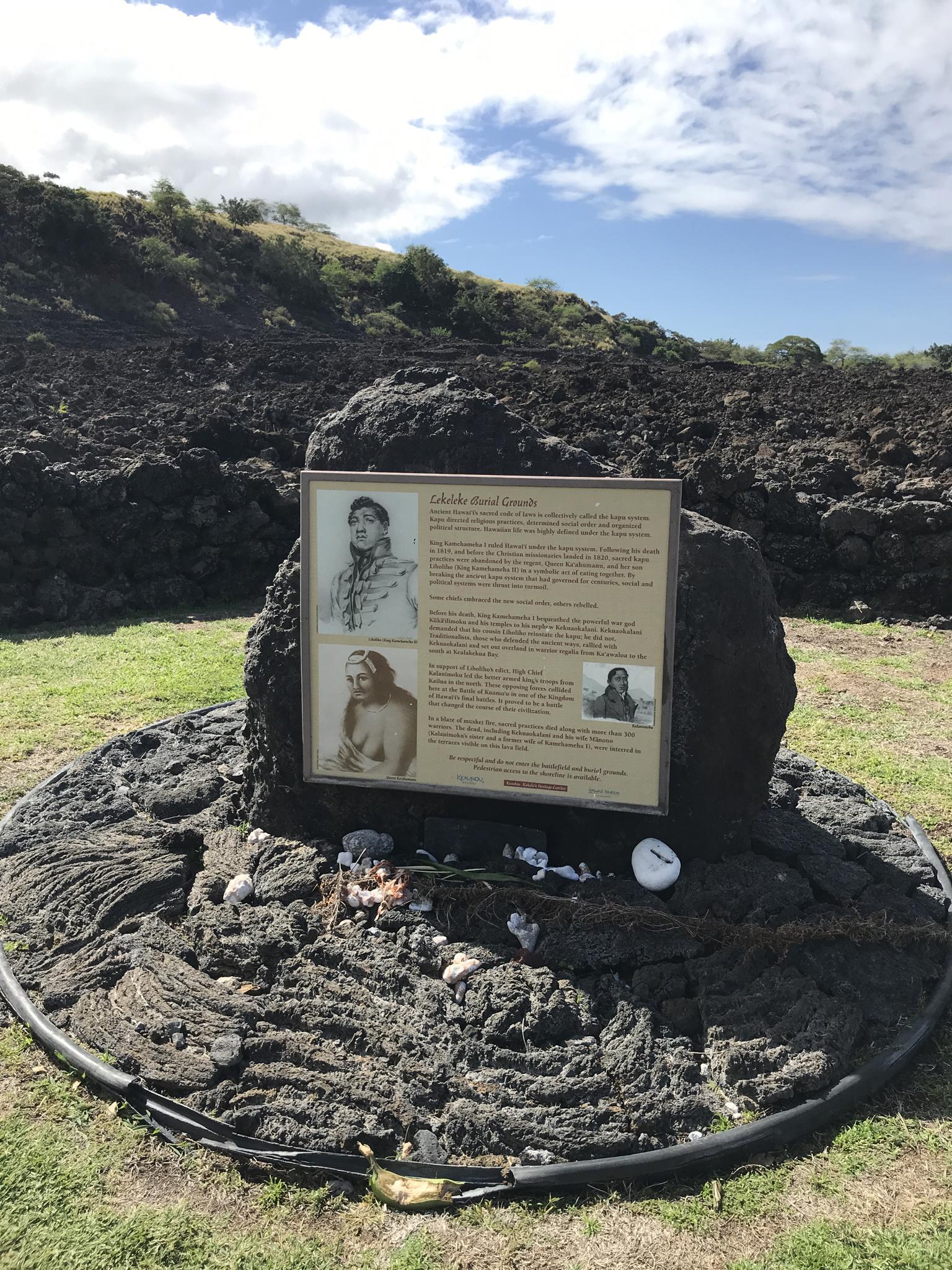 Memorial plaque on a large dark rock, green grass, and cloudy sky.