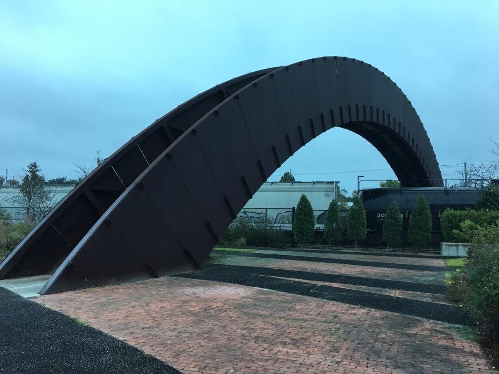 A large, dark, arched metal sculpture on paved ground under an overcast sky.