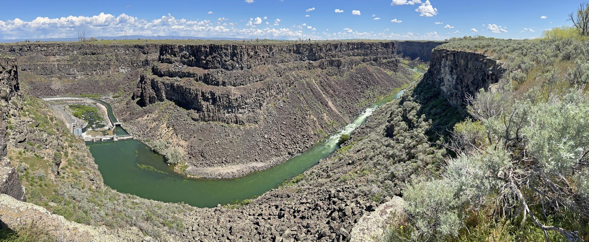 Wide canyon with a green river, dam, and rocky walls under a bright sky.