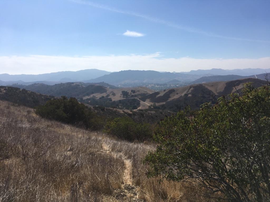 A narrow dirt path through dry grass and shrubs, with distant hills.