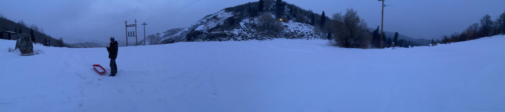 A person stands with a red sled on a snowy slope at dusk.