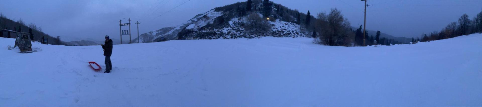 A person stands with a red sled on a snowy slope at dusk.