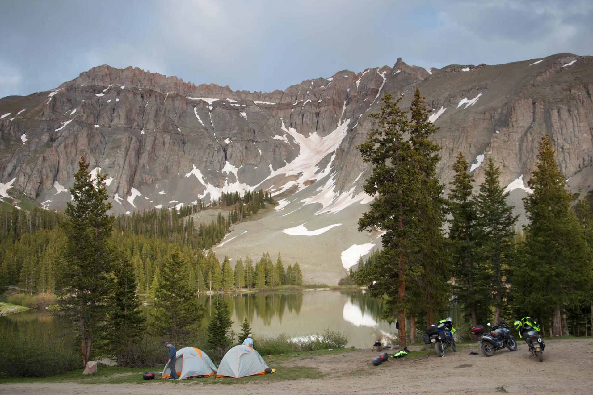 Tents and motorcycles by a lake with snowy mountains under an overcast sky.