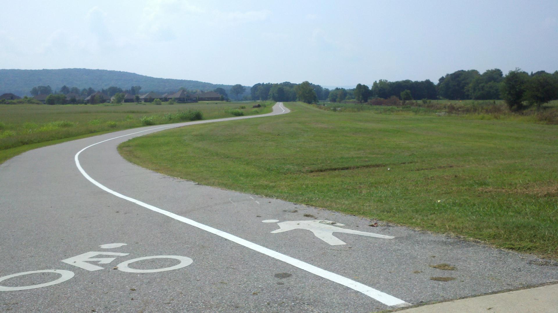 A paved multi-use path with bike and pedestrian symbols winds through green fields.