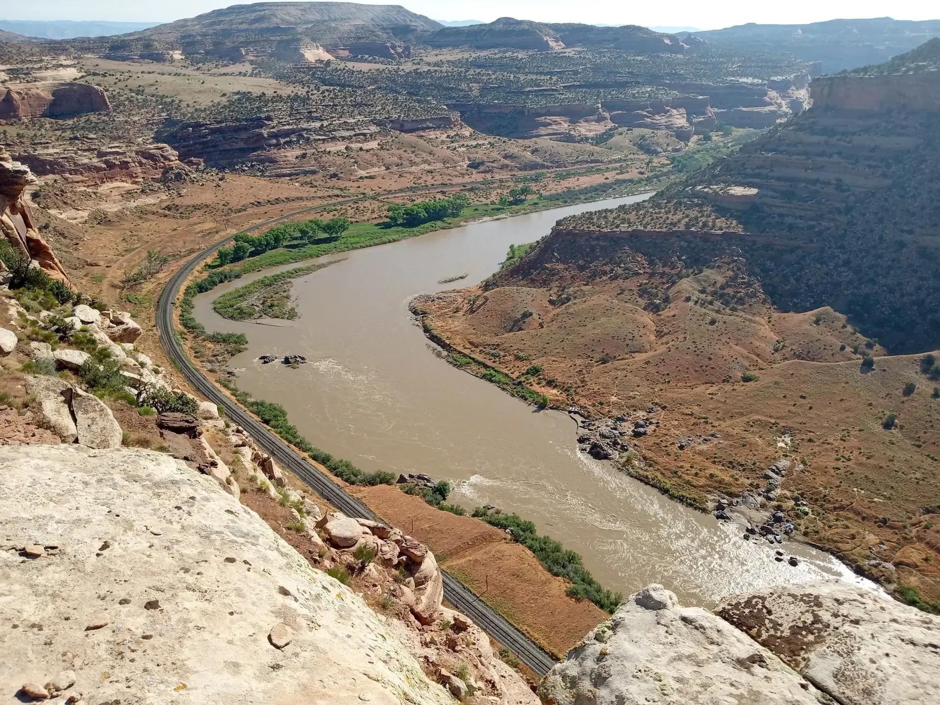 Winding river through an arid canyon with a railway track under bright sunlight.
