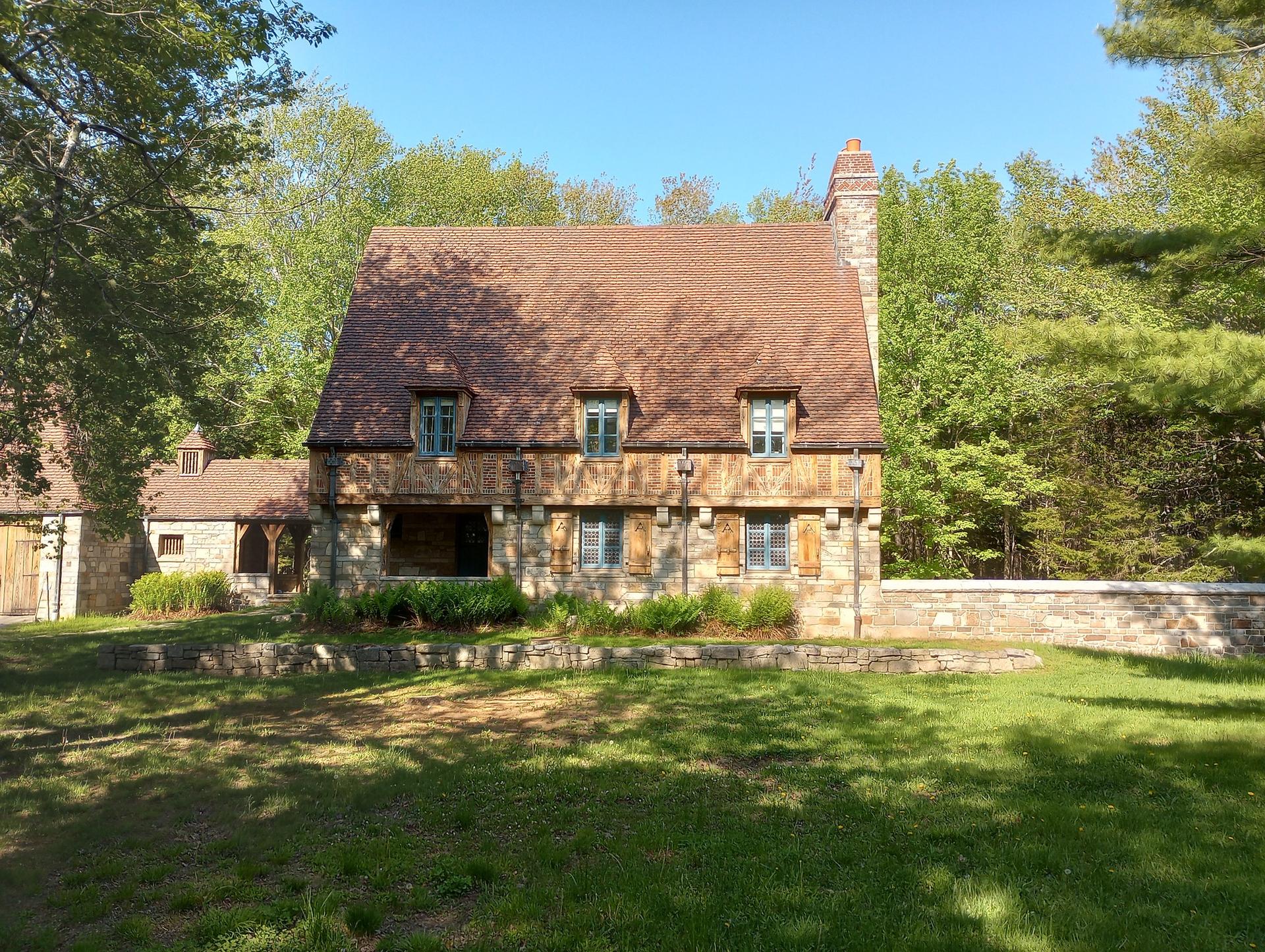 Rustic stone house with brown roof and dormers, surrounded by green trees on a sunny day.