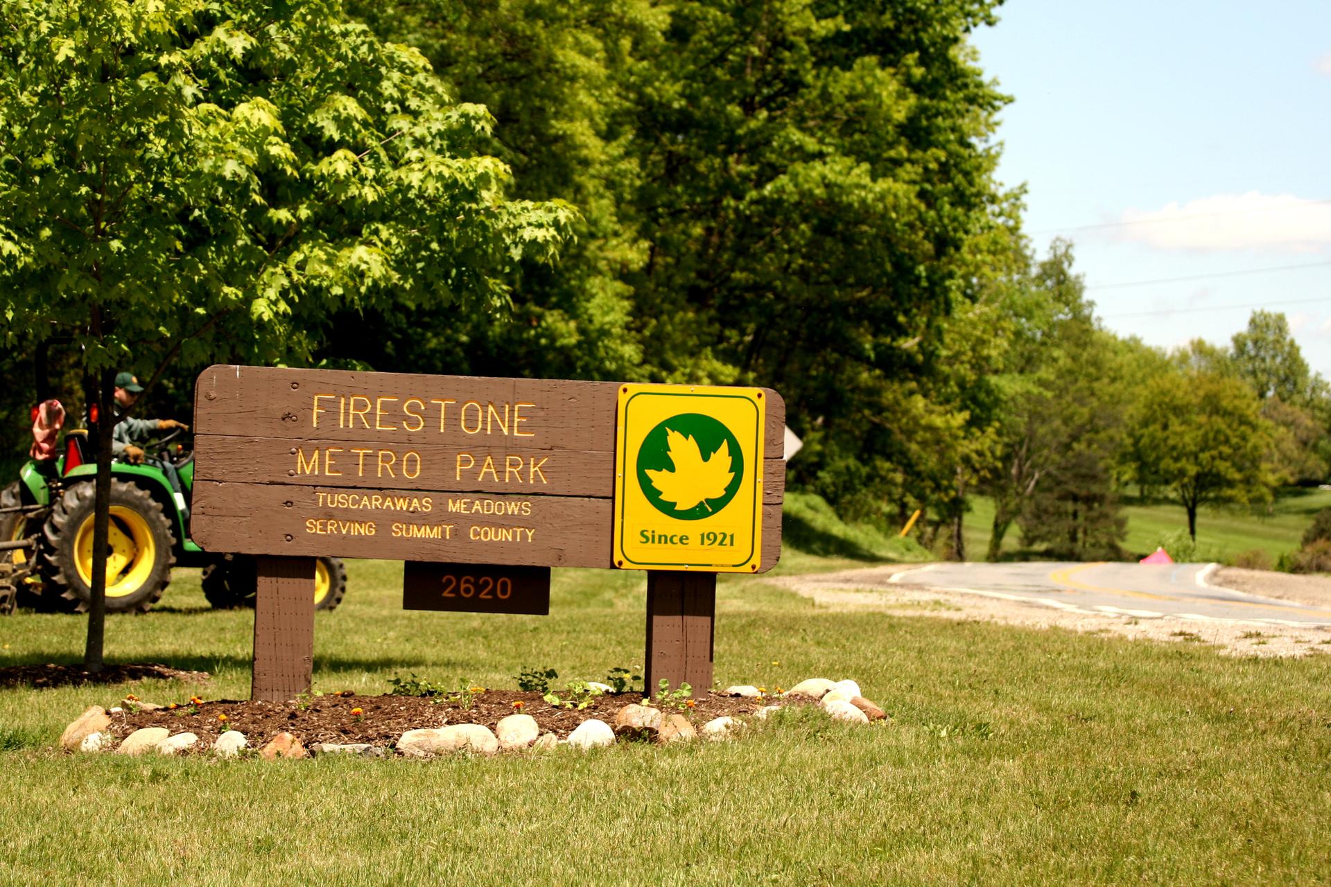 Sunny park entrance sign with a green tractor and lush trees.