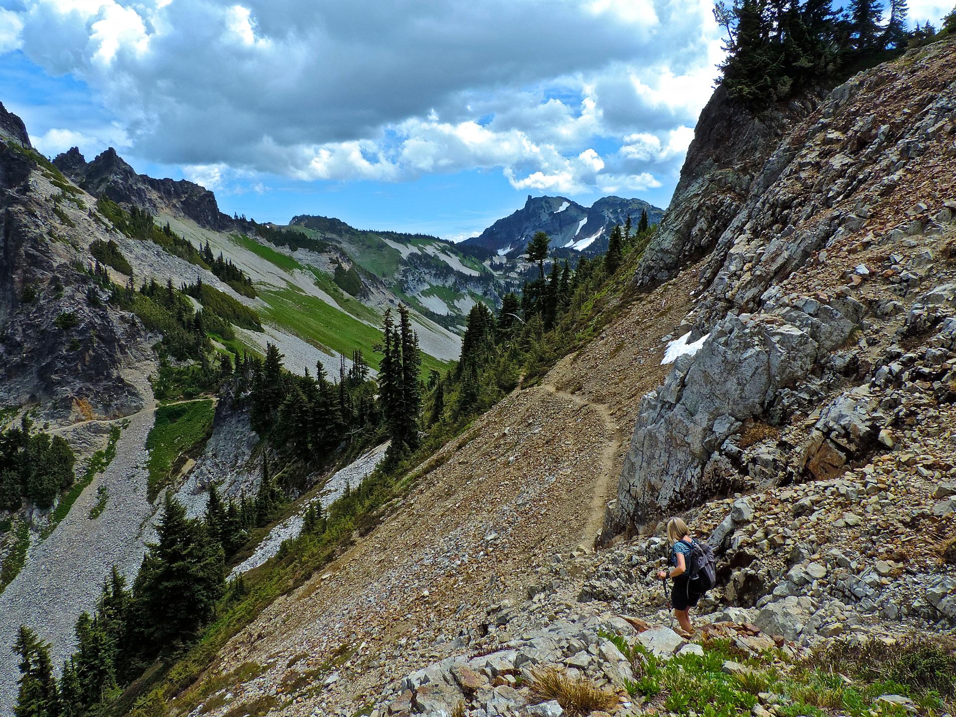 Hiker on a steep, rocky mountain trail overlooking a vast green valley.