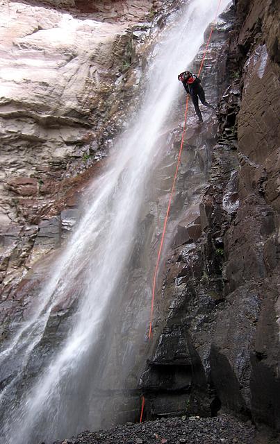 Person rappelling down a steep rock face beside a powerful waterfall.
